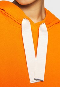 Hoodie arancione con una texture morbida, dotato di lacci bianchi piatti che mostrano testo in rilievo e estremità metalliche.