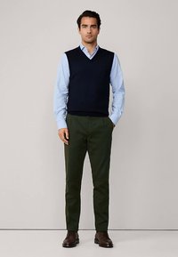 Laivaston sininen neuleliivi vaaleansinisen puseron päällä, yhdistetty tummanvihreisiin housuihin ja ruskeisiin nahkakenkiin, seisomassa neutraalilla taustalla.