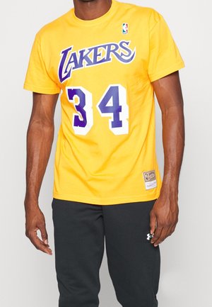 Mężczyzna w żółtej koszulce koszykarskiej Los Angeles Lakers z numerem 34 oraz czarnych spodniach Under Armour, stojący na jednolitym tle.