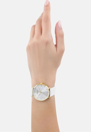 Weißer Uhr mit Gehäuse in Goldton und silbernem Ziffernblatt. Verfügt über minimalistische Stundenmarkierungen und ein glattes Lederarmband. Hand hochgehoben.