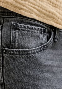 Hästi suumitud halli teksapükste tasku, kus on nähtavad õmblemised ja mustad naelad, kantud beeži tekstuurse kangaga.