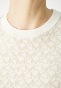 Svetr s krátkým rukávem v jemné krémové barvě, zdobený subtilním geometrickým vzorem v béžové. Jegovy detail kolem krku a měkká, strukturovaná tkanina.