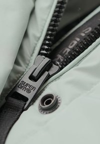 Lys grøn pufferjakke med en sort lynlås, der har et "SUPER DRY" logo. Inkluderer stoftekstur detaljer og en synlig trykknap.