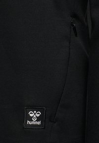 Black athletic pants featuring a zip pocket and a square logo patch with "hummel" text. Smooth fabric with a subtle texture.