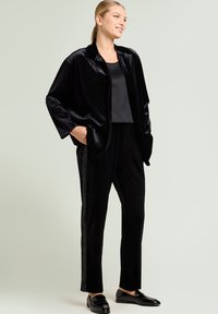 Black velvet jacket with wide sleeves and side pockets paired with matching trousers featuring a textured side stripe. Black loafers complete the outfit.
