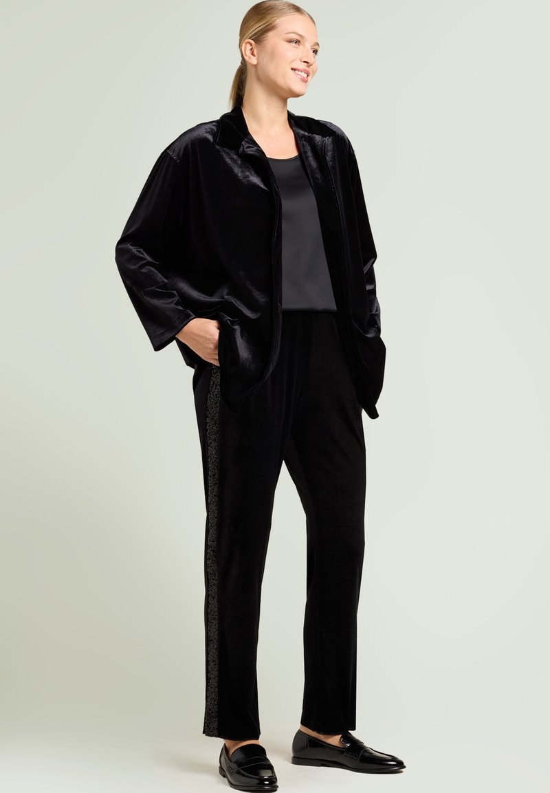 Black velvet jacket with wide sleeves and side pockets paired with matching trousers featuring a textured side stripe. Black loafers complete the outfit.