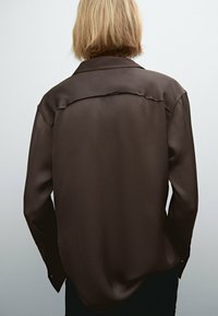Brun satin langærmet skjorte med klassisk krave, buet kant og subtil glans. Bagfra ses løs pasform og synlige sømdetaljer.