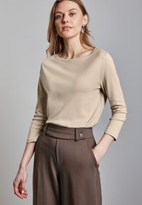 Beige Langarmshirt aus glattem Stoff, kombiniert mit hochgeschnittenen braunen Hosen mit einem Knopfdetail und seitlichen Taschen.