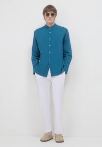 Camicia azzurro turchese con colletto alla mandarino, abbinata a pantaloni bianchi e scarpe slip-on beige; tessuto morbido e vestibilità rilassata.