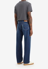 Camiseta de algodón gris oscuro combinada con jeans holgados de mezclilla azul. Los jeans tienen bolsillos traseros y una etiqueta de cuero marrón claro con la marca. Zapatillas blancas.