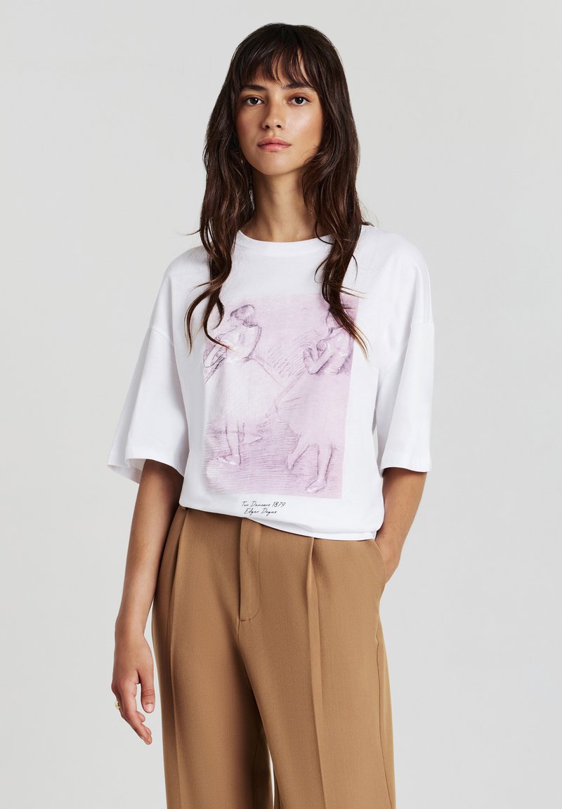 T-shirt bianco a maniche corte con una grafica rosa di ballerini. Il tessuto sembra morbido. Il T-shirt ha una vestibilità rilassata e un colletto rotondo, abbinato a pantaloni beige.