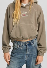 Harmaa fleecehuppari, jossa on nyöritys helmassa, edessä kengurutasku ja kirjailtu teksti. Yhdistetty korkeavyötäröisiin farkkuihin.