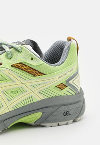 Gros plan d'une chaussure de course vert clair et grise avec une semelle texturée, une doublure en mesh et une marque "GEL-VENTURE 7" sur le côté.