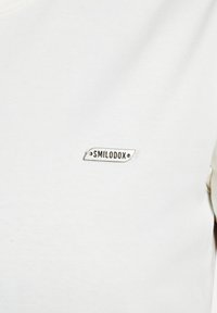 Hvid bomulds t-shirt med et lille rektangulært broderet mærke med teksten "SMILODOX." Stoffet har en glat, ensartet tekstur.