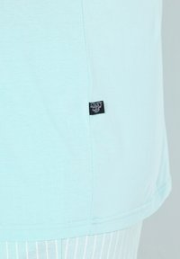 Camiseta de color azul claro, hecha de algodón suave y con un corte recto. Presenta una pequeña etiqueta de tela negra en el lado izquierdo, cerca del dobladillo.