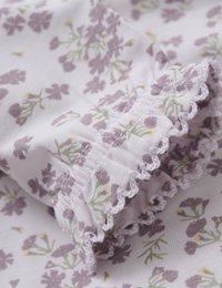 Fliederfarbener Stoff mit floralen Motiven, der kleine lila Blumen und grüne Stiele zeigt, mit gerafftem Saum und Spitzenelementen entlang des Randes.