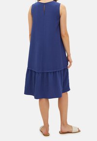 Sininen hihaton mekko, jossa on teksturoitu kangas, levenevä alaosa ja takana avonainen yksityiskohta. Yhdistetty beigen värisiin liukusandaaleihin.