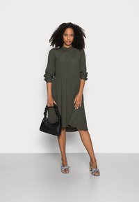 Vestido camisa verde oliva com fecho de botão, mangas longas e saia evasê. Combinado com uma bolsa preta e sandálias de salto com padrão.
