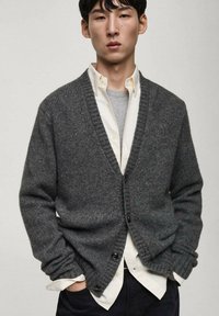 Šedý pletený cardigan s výstřihem do V, žebrovanými manžetami a zapínáním na knoflíky. Nesený přes světle šedé tričko a bílé košili.