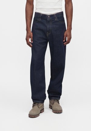 Homme portant un jean droit bleu foncé, des bottes marron clair à lacets et un débardeur blanc, debout devant un fond uni.