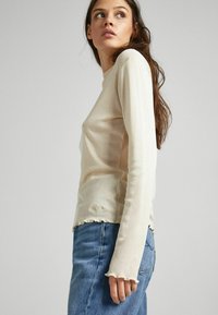 Maglietta a maniche lunghe di un beige chiaro con scollatura rotonda, caratterizzata da bordi sfilacciati e una vestibilità slim, abbinata a jeans blu a vita alta.