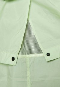 Tissu vert clair avec une ouverture d'aération triangulaire en mesh sécurisée par des boutons-pression noirs de chaque côté.
