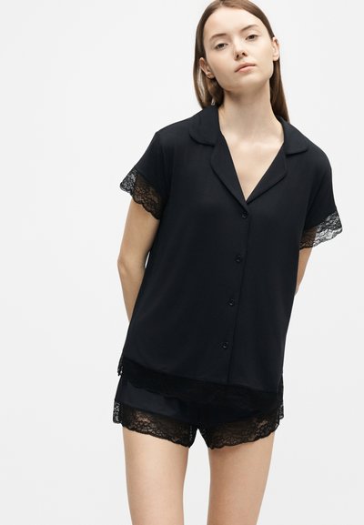 Jeune femme portant un haut noir à manches courtes boutonné et un short assorti avec bordure en dentelle, debout devant un fond clair uni.