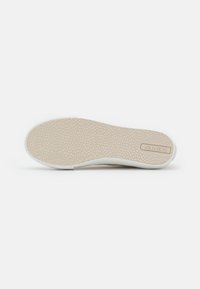 Semelle de sneaker blanche avec une surface en caoutchouc beige texturée, présentant un design plat et un logo subtil embossé sur le côté.
