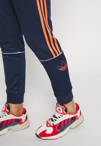 Námořnické sportovní tepláky s oranžovými bočními proužky a bílými detaily, s vyšitým logem na nohavici. V kombinaci s červenými a bílými teniskami.