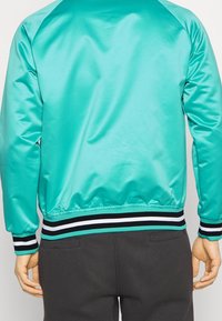 Chaqueta bomber sintética en color verde azulado con puños y dobladillo de canalé en blanco y negro, que presenta una textura suave y un ajuste holgado.