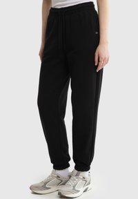 Persoon draagt zwarte joggers met elastische taille, zijzakken en witte sportschoenen, staand tegen een effen achtergrond.