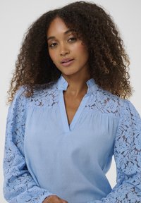 Blouse bleu clair avec détails en dentelle sur les manches, col mao et coupe ample avec un tissu subtilement texturé.