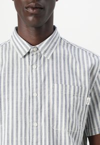 Camisa de botones de manga corta a rayas verticales en azul y blanco, hecha de algodón, con cuello y un bolsillo en el pecho. Presenta botones blancos.