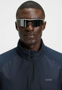Sonnenbrille mit elegantem Design, dunklen Gläsern und beigem Rahmen. Getragen mit einer dunkelblauen Jacke, die einen hohen Kragen und eine strukturierte Stoffqualität aufweist.