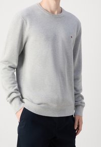 Grauer Strickpullover mit Rundhals und gerippten Bündchen. Verfügt über ein kleines Logo auf der Brust. Weicher Stoff, lässige Passform. Kombiniert mit dunklen Hosen.
