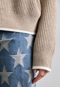 Beige ribbad tröja med vit kant över en jeanskjol prydd med stora vita stjärnor, där handen vilar vid sidan.
