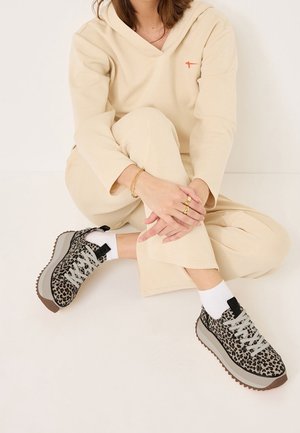 Personne portant un ensemble d’intérieur de couleur crème assise en tailleur, montrant des baskets compensées à imprimé léopard et des chaussettes blanches sur un fond clair.