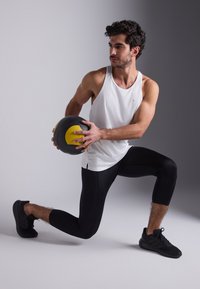 Weißes sportliches Tanktop, schwarze eng anliegende Capri-Leggings und schwarze Sneakers. Hält einen schwarz-gelben Medizinball in einer Ausfallschritt-Position.