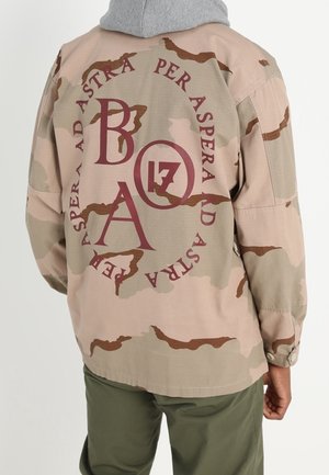 Osoba ubrana w beżowo-brązową kurtkę moro z czerwonym napisem "PER ASPERA AD ASTRA" i "B A 17" na plecach.