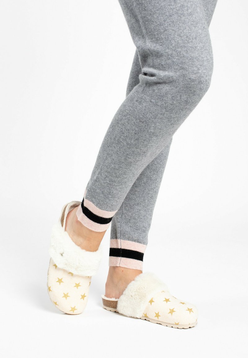 Weiße Clogs mit goldenen Sternmustern und einem flauschigen Kunstpelzkragen, kombiniert mit grauen Jogginghosen, die an den Knöcheln schwarze und rosa Streifen haben.