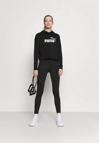Μαύρο cropped φούτερ με λευκό λογότυπο Puma, συνδυασμένο με μαύρο κολάν. Το μοντέλο κρατά μαύρο όργανο γυμναστικής, φορώντας λευκά αθλητικά παπούτσια.