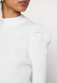 Pull blanc côtelé avec un col montant et des manches bouffantes texturées. Le tissu semble doux et légèrement extensible. Design épuré et minimaliste.