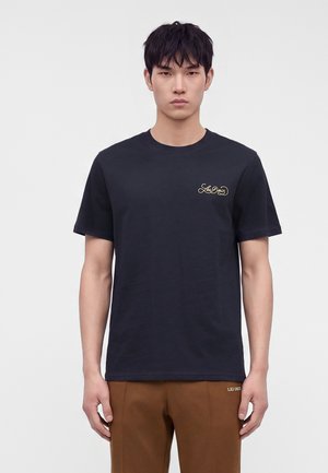 Νέος με κοντά μαύρα μαλλιά που φοράει μαύρο T-shirt με μικρό κίτρινο λογότυπο και καφέ παντελόνι σε απλό λευκό φόντο.