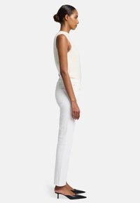 Kremowy bezrękawnik, dopasowane białe jeansy i czarne szpilki. Gładki materiał, dopasowany krój i wyraźna sylwetka. Widok z boku.