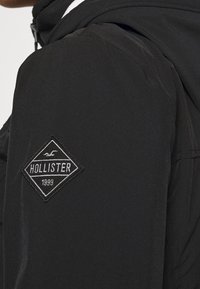 Svart jacka med en slät yta och huva, med en diamantformad broderad logotyp som läser "Hollister 1999" på ärmen.