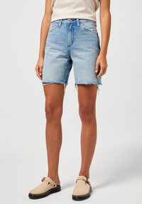 Shorts in denim azzurro chiaro con vita alta, orlo sfilacciato e due tasche frontali. Indossati con scarpe beige senza lacci con suola nera.