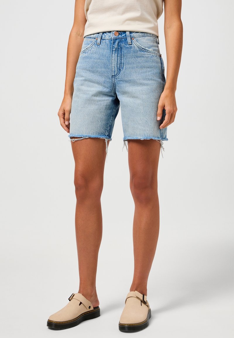 Shorts in denim azzurro chiaro con vita alta, orlo sfilacciato e due tasche frontali. Indossati con scarpe beige senza lacci con suola nera.