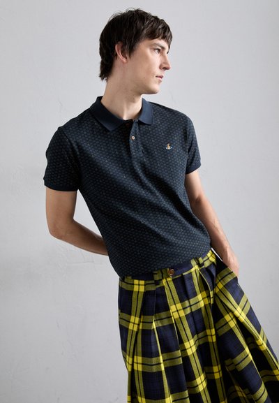 Dunkelblaues Poloshirt mit geometrischem Muster, kombiniert mit einem gelb- und marineblauen Karokilt, der Falten und ein Knopfdetail an der Taille aufweist.