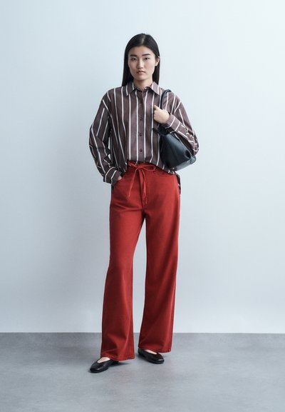 Γυναίκα που στέκεται φορώντας πουκάμισο με καφέ και λευκές ρίγες, κόκκινο παντελόνι με φαρδιά μπατζάκια, μαύρες μπαλαρίνες και κρατώντας μια μαύρη τσάντα ώμου.