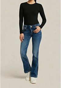 Personne portant un haut noir à manches longues, un jean évasé bleu et des chaussures blanches, debout avec une main posée sur la hanche et l'autre le long du corps.